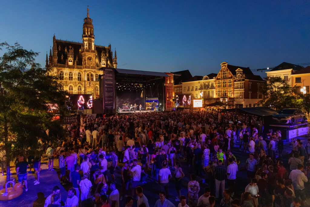 Avondbeeld van een vol Marktplein tijdens de Bierfeesten in Oudenaarde, met een optreden op het hoofdpodium en het verlichte stadhuis op de achtergrond.